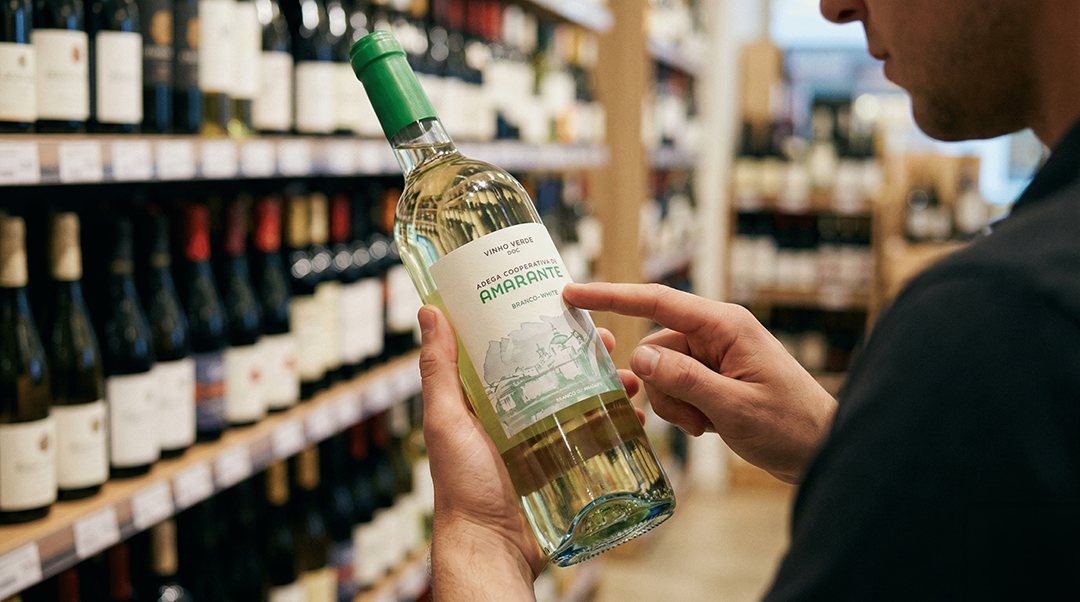 Close-up de um comprador segurando e analisando o rótulo de uma garrafa de vinho branco, com prateleiras de vinho ao fundo